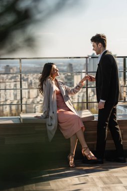 Elbiseli ve ceketli mutlu bir kadın çatıda takım elbiseli bir adamla el ele tutuşuyor.