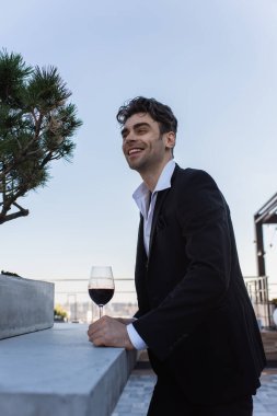 Takım elbiseli neşeli adam terasta kırmızı şarap tutuyor. 