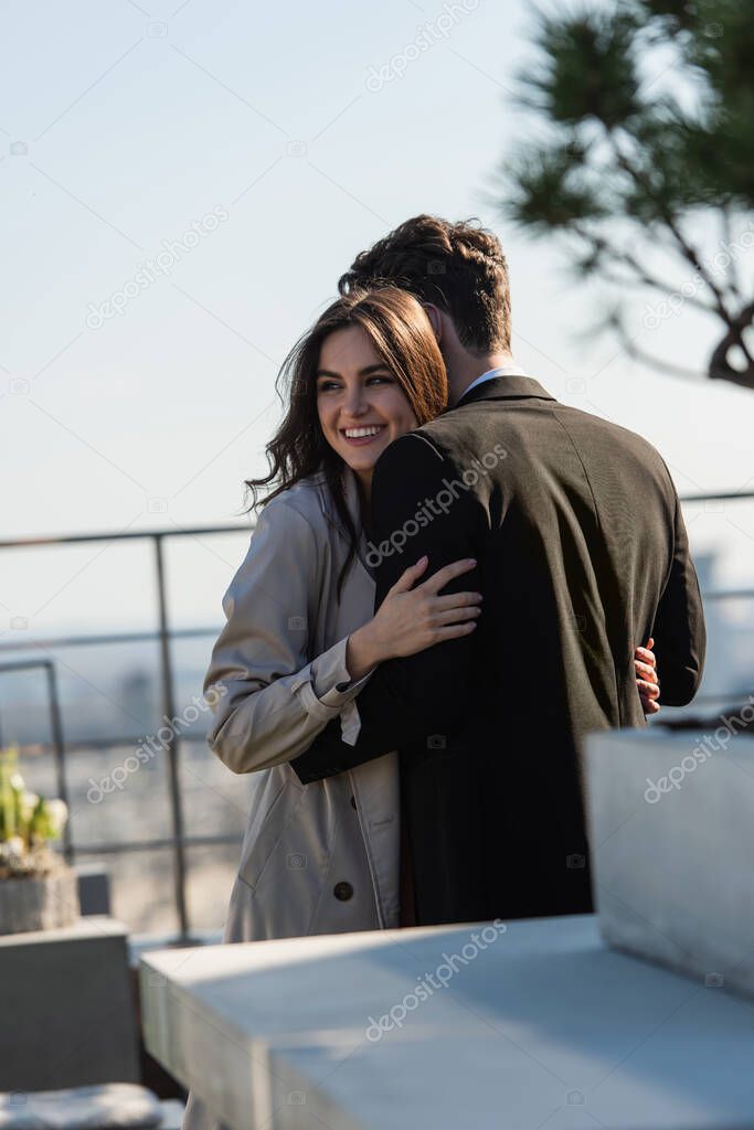 man hugging cheerful woman in coat on terrace 