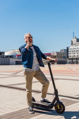 full length of happy and mature man in sunglasses talking on smartphone while standing with e-scooter outside 