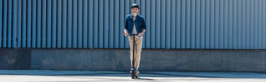 full length of mature man in helmet and sunglasses riding electric scooter on street, banner