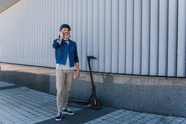 full length of cheerful and mature hipster in helmet and sunglasses talking on smartphone near e-scooter 