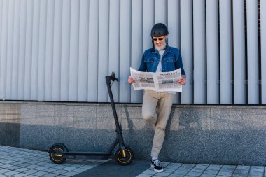 full length of happy and mature hipster in helmet and sunglasses reading travel life newspaper near e-scooter 