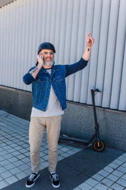full length of cheerful and mature hipster in helmet and sunglasses talking on smartphone and gesturing near e-scooter 
