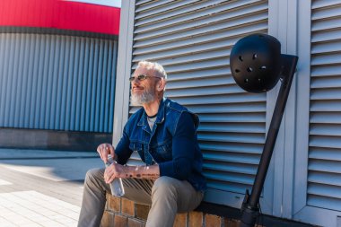 mature man in sunglasses smiling and holding bottle of water near e-scooter with helmet