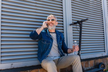 cheerful and mature man in sunglasses talking on smartphone and holding bottle of water near e-scooter 