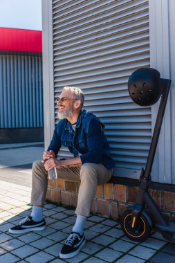 bearded mature man in sunglasses smiling and holding bottle of water near e-scooter with helmet