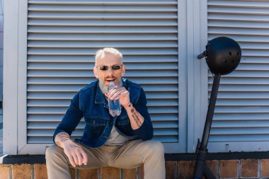 bearded mature man in sunglasses smiling and drinking water near e-scooter with helmet
