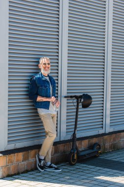 bearded mature man in sunglasses smiling and holding bottle of water while standing near e-scooter with helmet