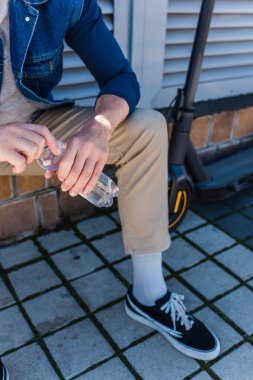 cropped view of mature man sitting and holding bottle of water near e-scooter 
