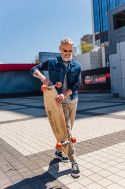 tattooed and middle aged man in sunglasses holding longboard on urban street