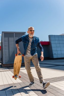 focused middle aged man in sunglasses holding longboard on urban street