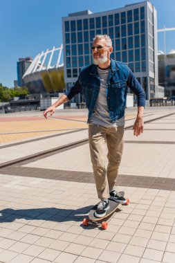 joyful middle aged man in sunglasses riding longboard on urban street