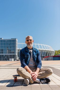 positive middle aged man in sunglasses sitting on longboard outside 