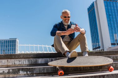 happy middle aged man in sunglasses sitting on stairs near longboard while using cellphone on urban street
