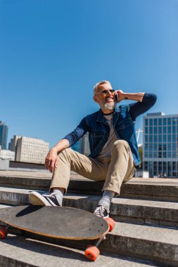 happy middle aged man in sunglasses sitting on stairs near longboard while talking on cellphone on urban street