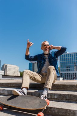 excited middle aged man in sunglasses sitting on stairs near longboard while talking on cellphone and showing rock sign on urban street