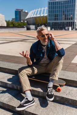 happy mature man in sunglasses sitting on stairs near longboard while talking on cellphone on urban street