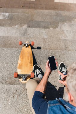 top view of tattooed middle aged man using smartphone with blank screen near longboard on stairs 