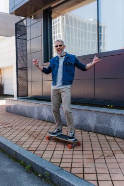 full length of excited man with open mouth riding longboard and showing rock sign on urban street