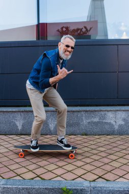 full length of happy man with open mouth riding longboard and showing rock sign on urban street