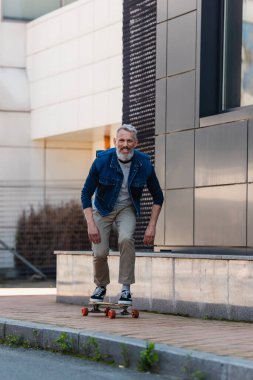 full length of middle aged man smiling and riding longboard on urban street