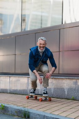 full length of cheerful and middle aged man riding longboard on urban street