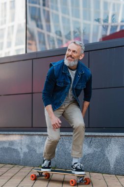 full length of smiling middle aged man riding longboard on urban street