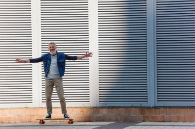full length of tattooed and happy middle aged man with outstretched hands riding longboard on urban street