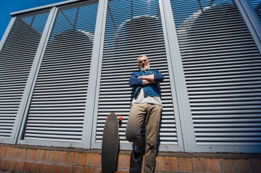 low angle view of positive and middle aged man standing with crossed arms near longboard on street