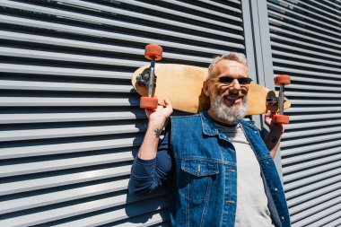 bearded middle aged man in sunglasses smiling and holding longboard 