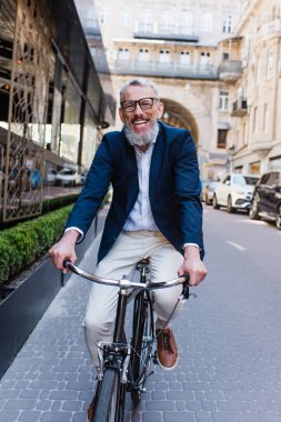 pleased middle aged man in glasses riding bicycle on modern urban street 