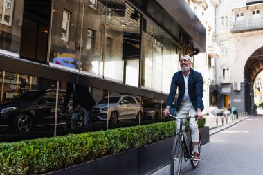 smiling middle aged man in blazer and glasses riding bicycle on modern urban street 