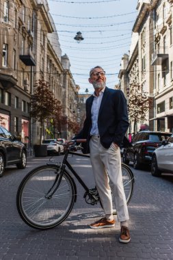happy middle aged man in blazer and glasses standing with hand in pocket near bicycle on modern urban street 