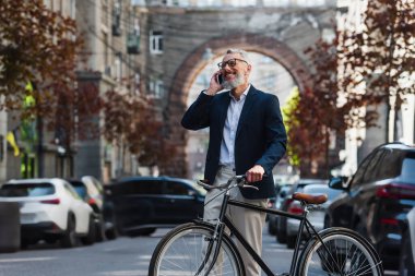 joyful middle aged man in blazer and glasses talking on smartphone and standing near bicycle on modern urban street 