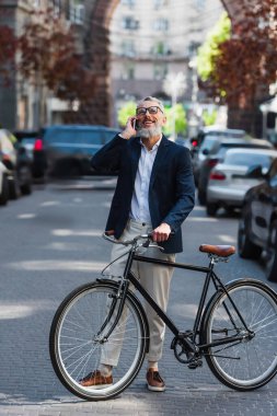 smiling middle aged man in blazer and glasses talking on smartphone and standing near bicycle on modern urban street 