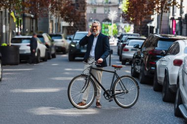 full length of happy middle aged man in blazer and glasses talking on smartphone and standing near bicycle on modern urban street 