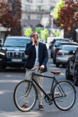 full length of middle aged man in blazer and glasses talking on smartphone and standing near bicycle on modern urban street 