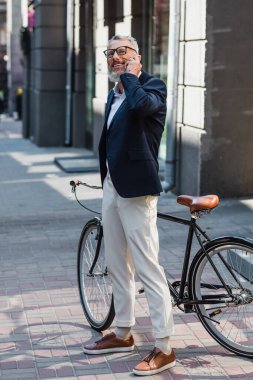 full length of cheerful middle aged man in blazer and glasses talking on smartphone and standing near bicycle on street 