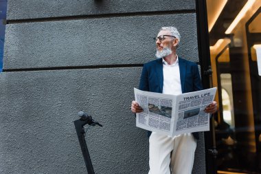 mature businessman in blazer standing with travel life newspaper near e-scooter 