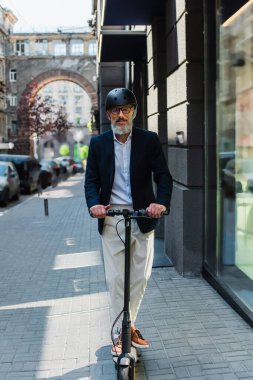 mature businessman in blazer and helmet riding e-scooter 