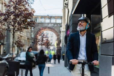 mature businessman in blazer and helmet riding e-scooter and looking away outside
