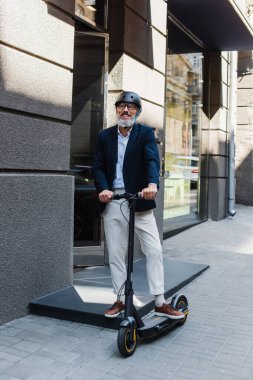 full length of happy and mature businessman in blazer and helmet riding electric scooter 