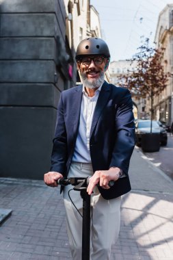 positive and mature businessman in blazer and helmet riding electric scooter 