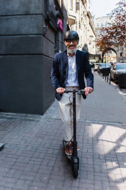 full length of positive and mature businessman in blazer and helmet riding electric scooter 