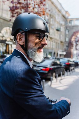 smiling and mature businessman in blazer and helmet riding electric scooter 
