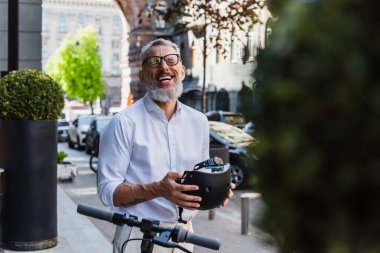 smiling and mature man in shirt and glasses laughing while holding helmet near electric scooter