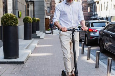 cropped view of tattooed and mature man in shirt riding electric scooter