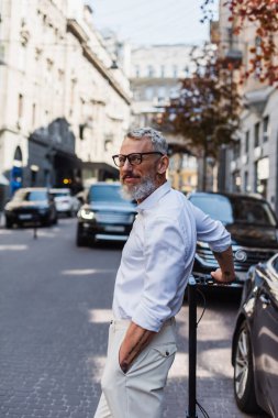 smiling and tattooed man in shirt standing with hand in pocket near electric scooter on street