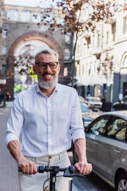 smiling and tattooed man in shirt riding e-scooter on street